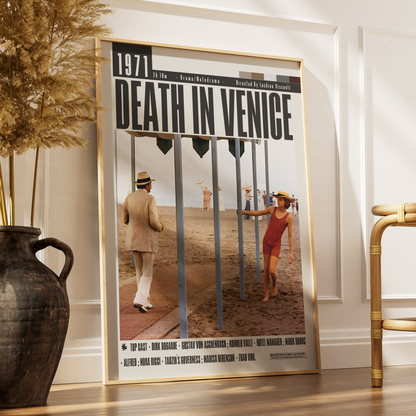 A framed vintage-style poster for the film 'Death in Venice' leaning against a white wall on a wooden floor, with a large dark vase and dried grass on the left and part of a gold chair on the right.