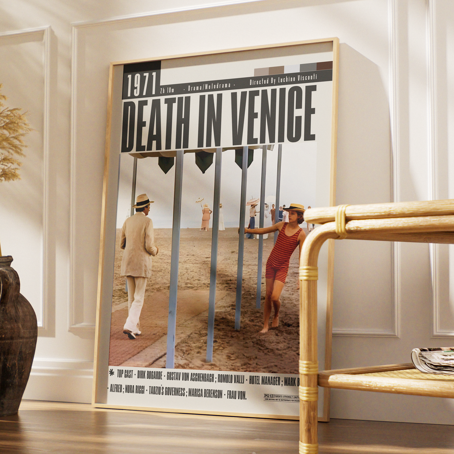 A framed poster of the movie 'Death in Venice' leaning against a white paneled wall on a wooden floor, next to a bamboo side table.