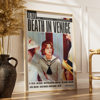 A framed 'Death in Venice' movie poster leaning against a white wall on a wooden floor, styled in a home interior next to a dark ceramic vase with dried grass and a bamboo chair.