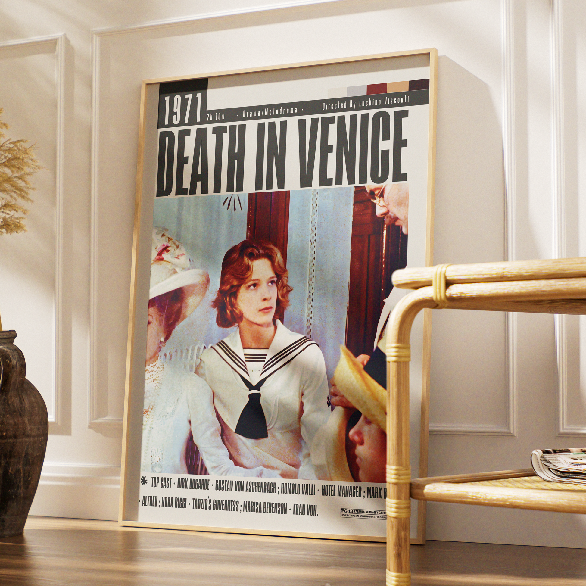 A framed vintage-style movie poster for 'Death in Venice' leaning against a white paneled wall on a wooden floor, with a bamboo side table partially visible in the foreground.