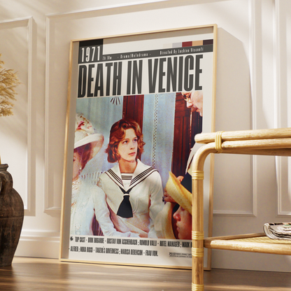 A framed vintage-style movie poster for 'Death in Venice' leaning against a white paneled wall on a wooden floor, with a bamboo side table partially visible in the foreground.