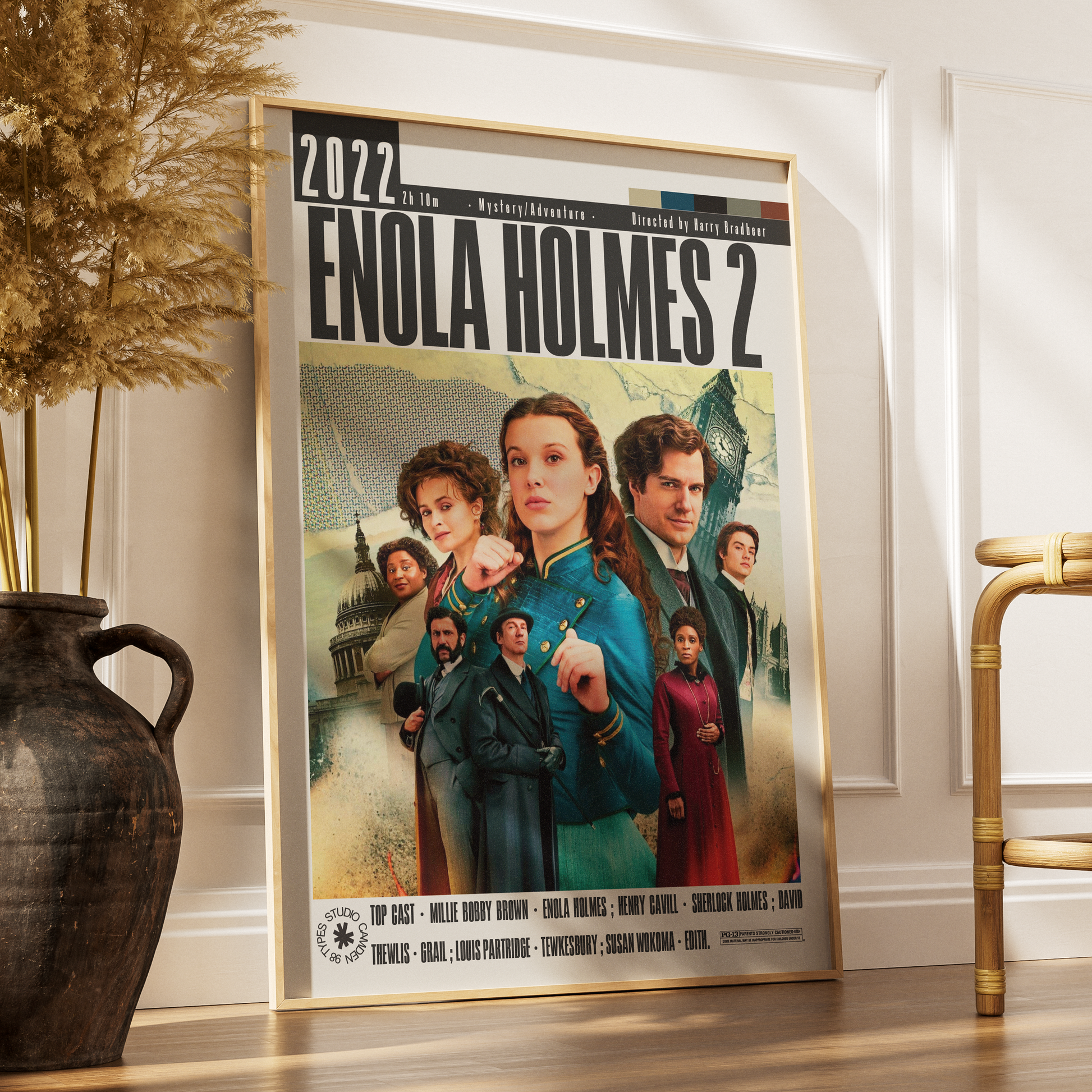 A framed movie poster for 'Enola Holmes 2' leaning against a white wall on a wooden floor, styled in a room with a dark vase of dried grass on the left and a gold chair on the right.