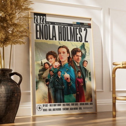 A framed movie poster for 'Enola Holmes 2' leaning against a white wall on a wooden floor, styled in a room with a dark vase of dried grass on the left and a gold chair on the right.