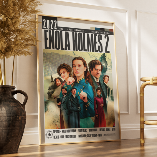 A framed movie poster for 'Enola Holmes 2' leaning against a white wall on a wooden floor, styled in a room with a dark vase of dried grass on the left and a gold chair on the right.