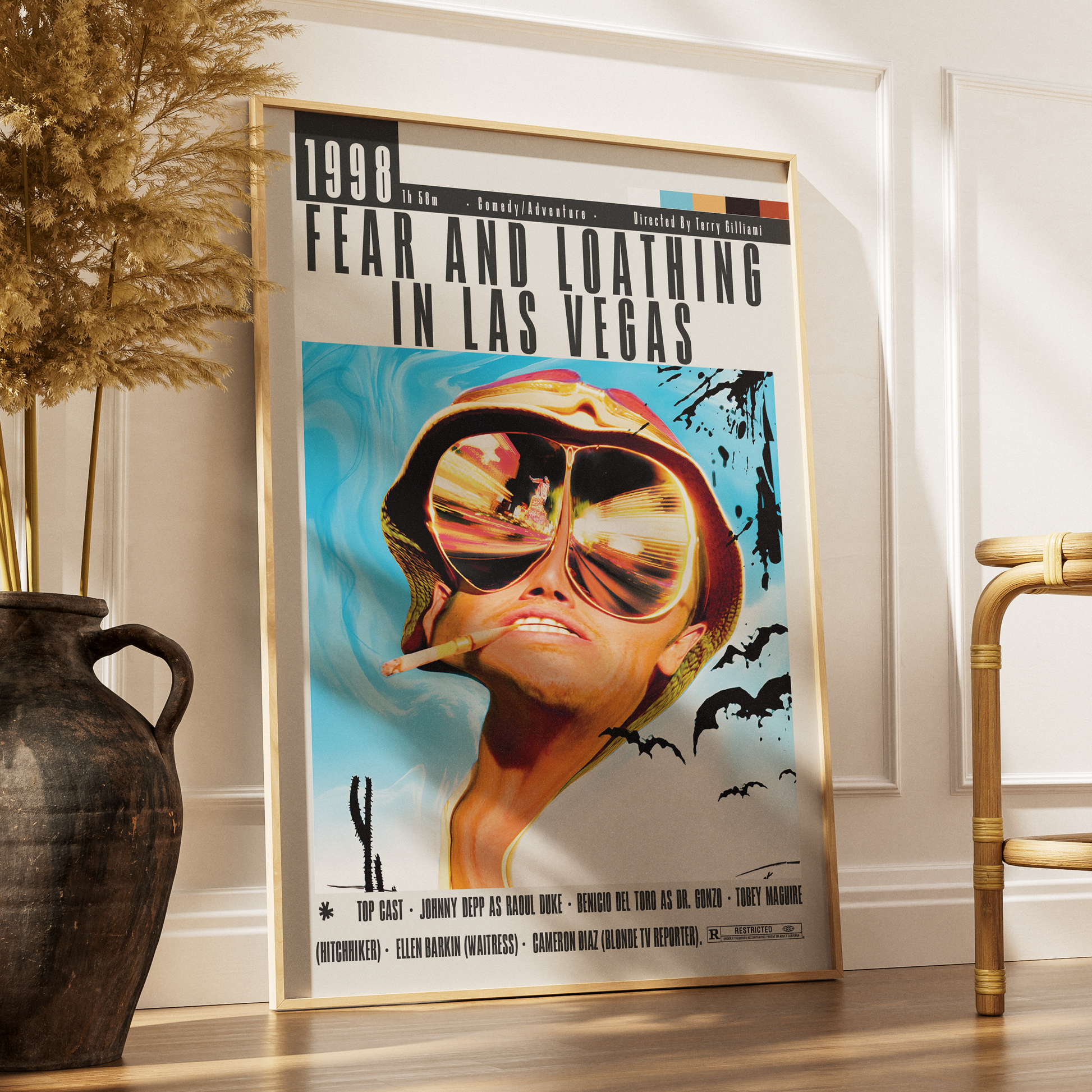 A framed vintage-style movie poster for 'Fear and Loathing in Las Vegas' leaning against a white wall on a wooden floor, styled in a room with a dark vase of dried grass on the left and a gold chair on the right.