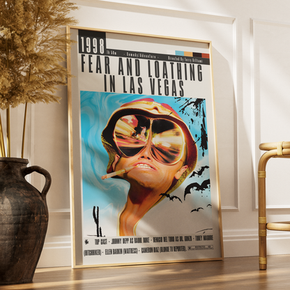 A framed vintage-style movie poster for 'Fear and Loathing in Las Vegas' leaning against a white wall on a wooden floor, styled in a room with a dark vase of dried grass on the left and a gold chair on the right.