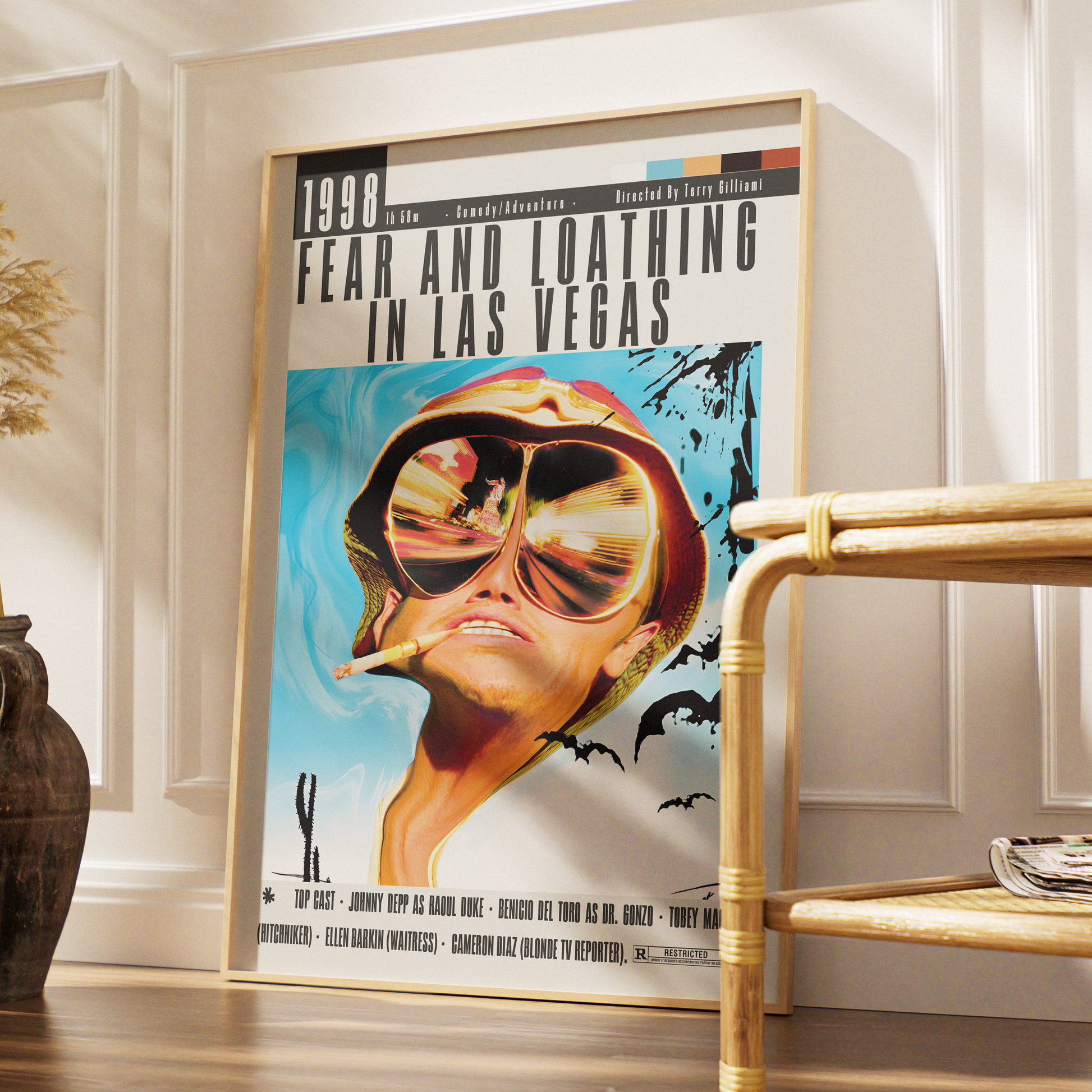 A framed vintage-style poster leaning against a white paneled wall in a styled interior. The poster reads '1998 FEAR AND LOATHING IN LAS VEGAS' and features a surreal illustrated portrait with oversized reflective sunglasses, a cigarette, bats, and a blue sky background.