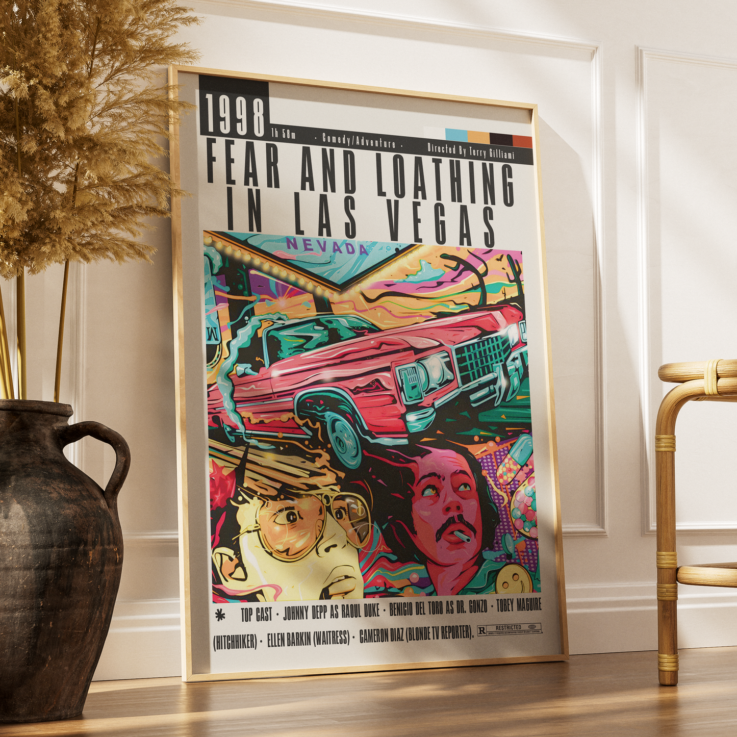 A framed pop art poster of the 1998 film 'Fear and Loathing in Las Vegas' leaning against a white wall on a wooden floor, styled in a room with a dark vase of dried grass on the left and a gold chair on the right.