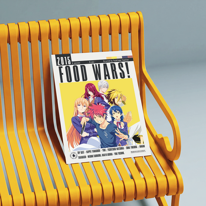 A printed magazine or book cover titled 'FOOD WARS!' with anime-style characters and a yellow cover, leaning on a bright yellow slatted bench against a soft gray studio background.