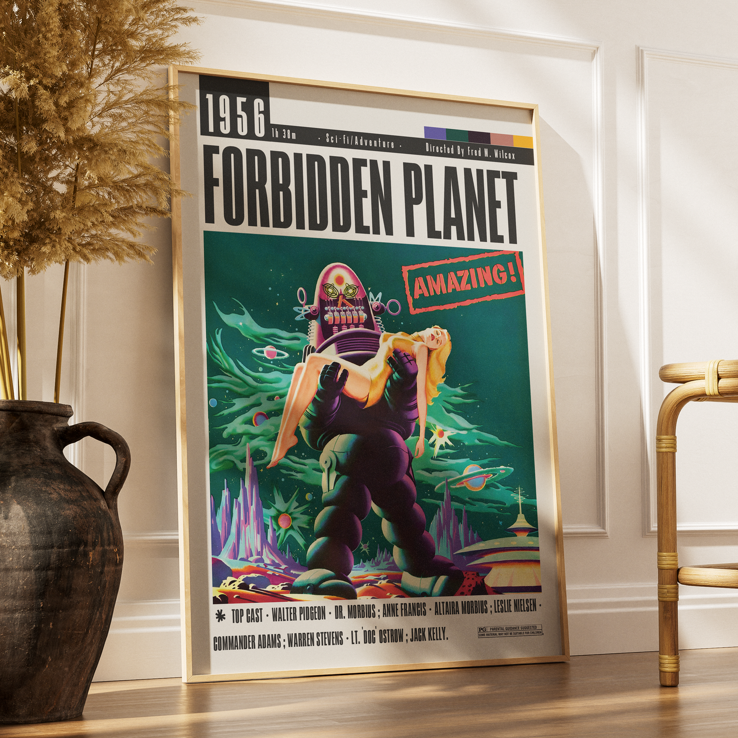 A framed retro-style poster for the movie 'Forbidden Planet' leaning against a white wall on a wooden floor, next to a dark ceramic vase with dried grass and part of a gold chair.