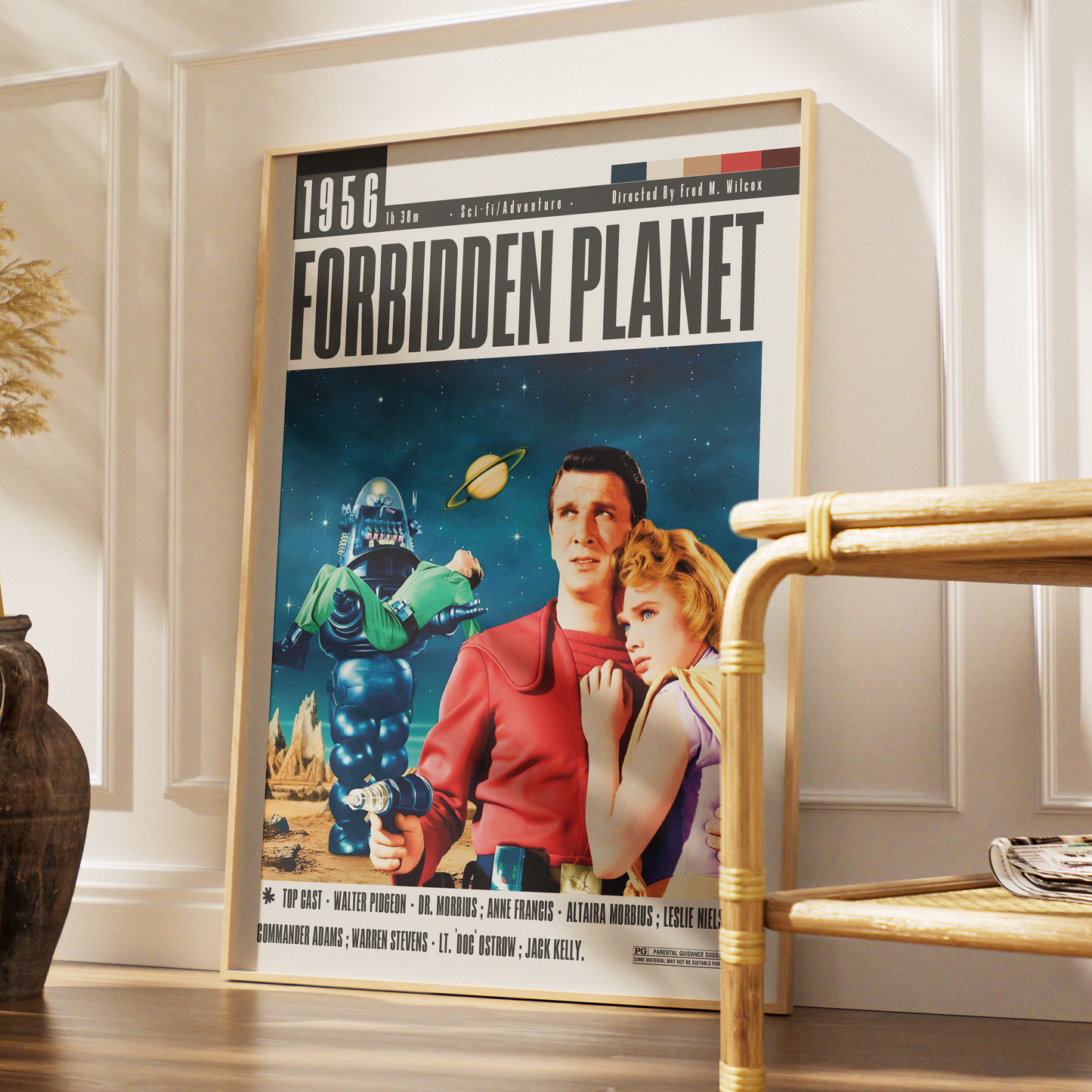 A framed vintage-style poster reading '1956 Forbidden Planet' leaning against a white paneled wall on a wooden floor, with a woven chair and decorative vase nearby.