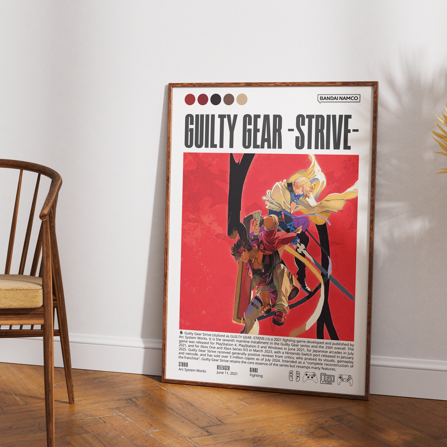 A framed 'Guilty Gear - Strive' poster leaning against a white wall on a wooden floor, with part of a wooden chair visible on the left. The poster features a red background, character artwork, and text including the 'Random Access' label.