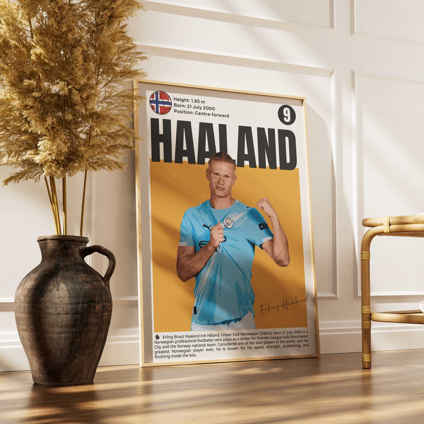 A framed sports poster of footballer Erik Haaland leaning against a white paneled wall on a wooden floor, styled with a dark ceramic vase of dried grass on the left and a gold-framed chair on the right.