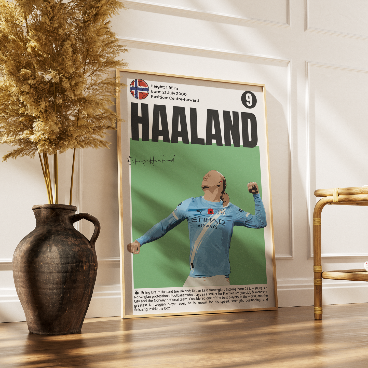 A framed sports poster of cyclist Jumbo Haaland leaning against a white paneled wall on a wooden floor, styled with a dark ceramic vase of dried grass on the left and a gold chair on the right.