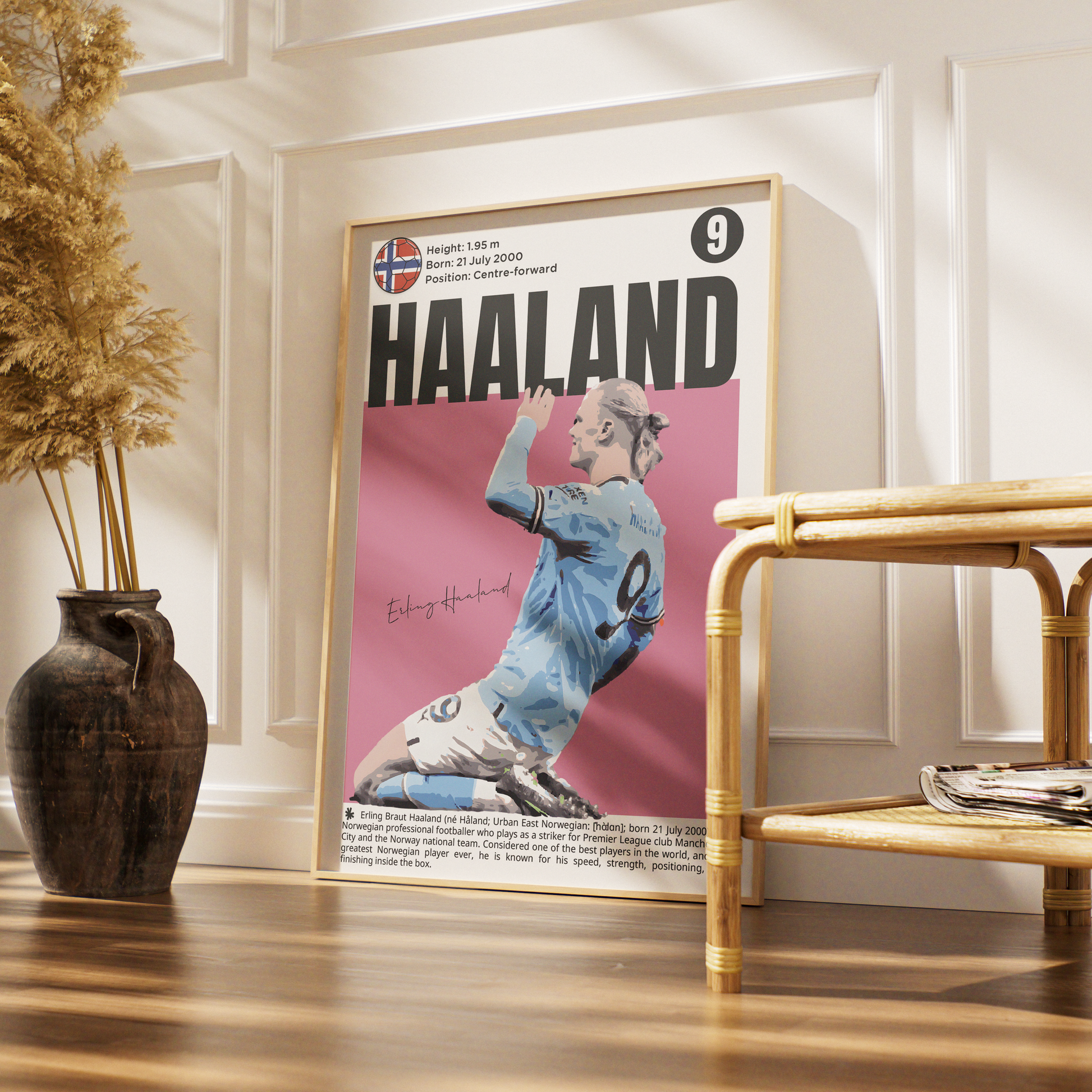 A framed poster of footballer Edin Dzeko 'Haaland' displayed on a wall in a styled room setting with a wooden floor, a rattan side table, and a dark ceramic vase with dried grass.