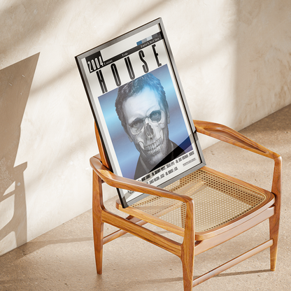A framed art print featuring a stylized portrait of a skull in a blue-toned magazine cover design, displayed on a mid-century modern wooden armchair with a woven cane seat in a sunlit room.