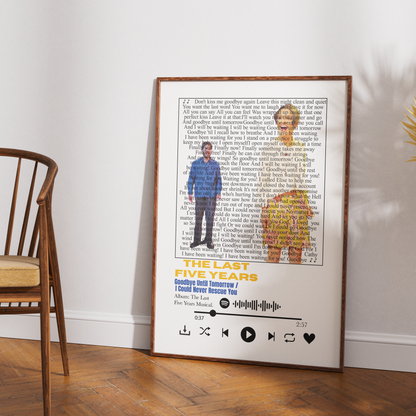 A framed music-themed wall print leaning against a white wall on a wooden floor, showing song lyrics, a photo of a man in a blue suit, a photo of a woman in a yellow dress, and playback icons at the bottom. A wooden chair is visible on the left.
