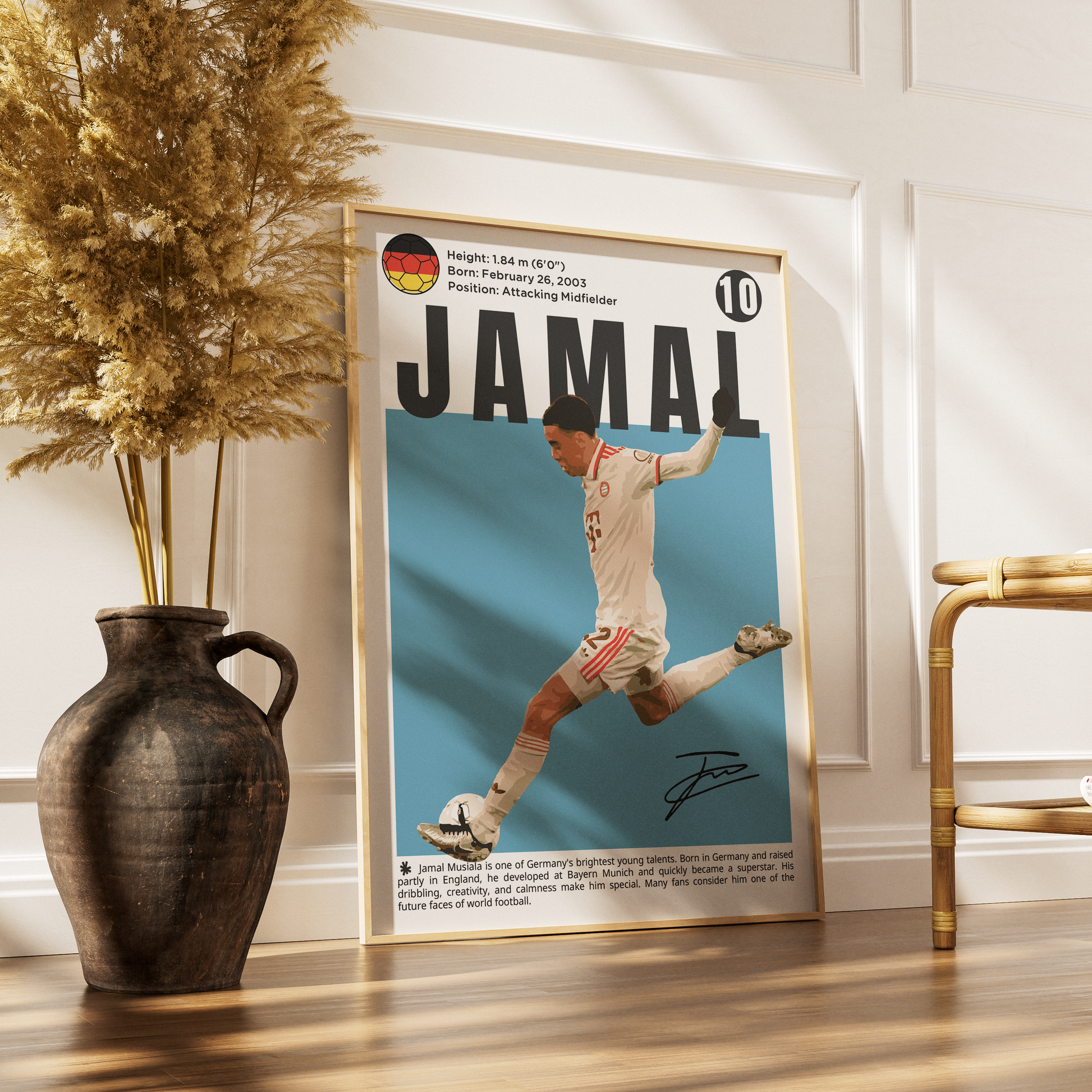A framed poster featuring a soccer player named Jamal on a light blue background, leaning against a white paneled wall on a wooden floor next to a dark vase with dried grass and a gold chair.