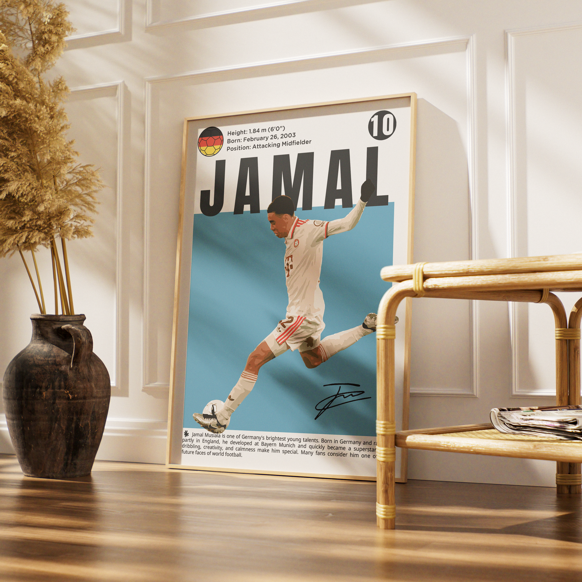 A framed vertical poster featuring a football player with the name JAMAL and a light blue background, leaning against a white paneled wall on a wooden floor next to a rattan bench and a dark vase with dried grass.