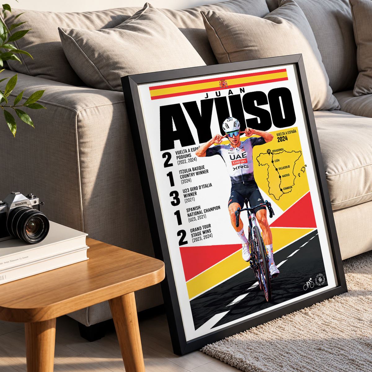 A framed cycling poster leaning against a beige sofa in a living room setting. The poster features Juan Ayuso with a large 'AYUSO' title, race statistics, a map of Spain, and a photo of the cyclist on a bicycle.