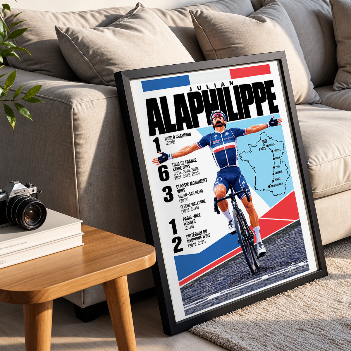 A framed cycling poster featuring Julian Alaphippe leaning against a beige sofa in a living room setting, with a small wooden side table and camera visible nearby.