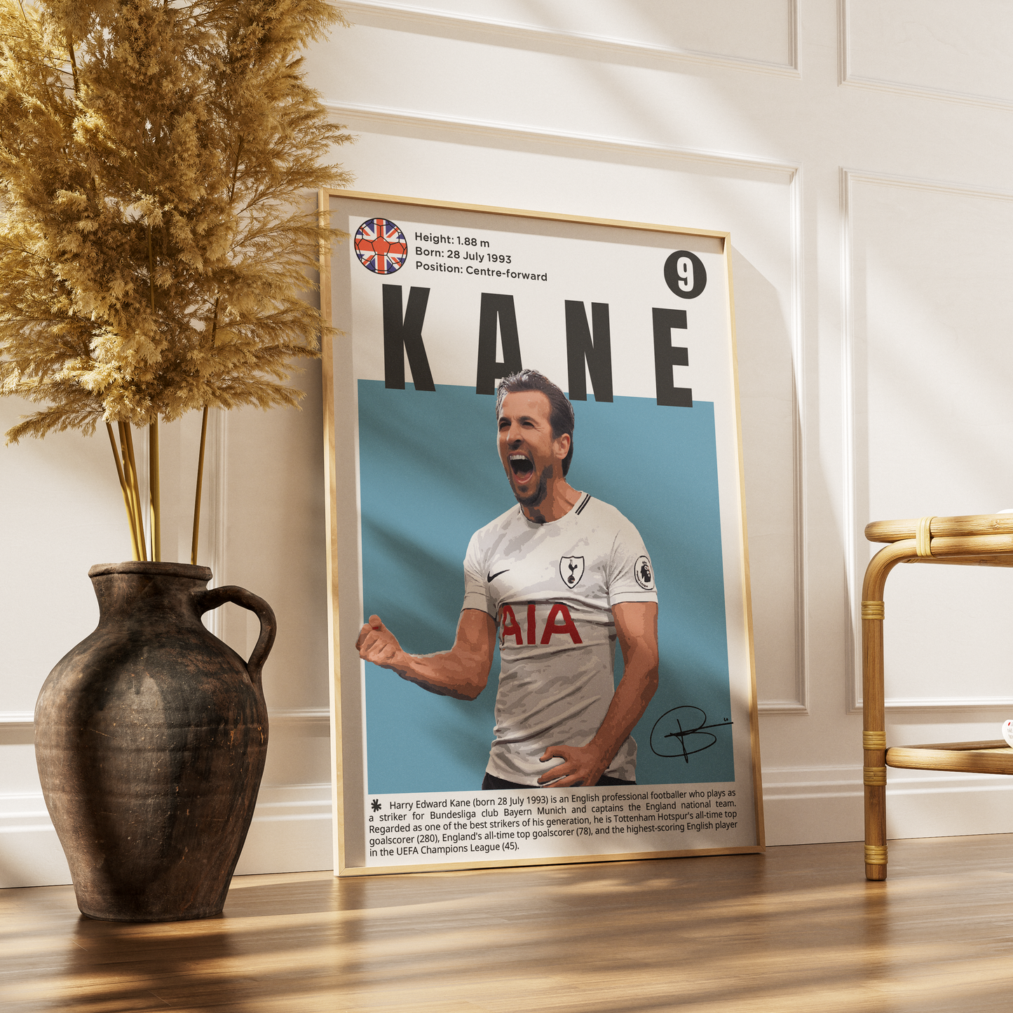 A framed vertical poster of footballer Harry Kane leaning against a white paneled wall on a wooden floor, styled with a dark ceramic vase of dried grass on the left and a gold chair on the right.