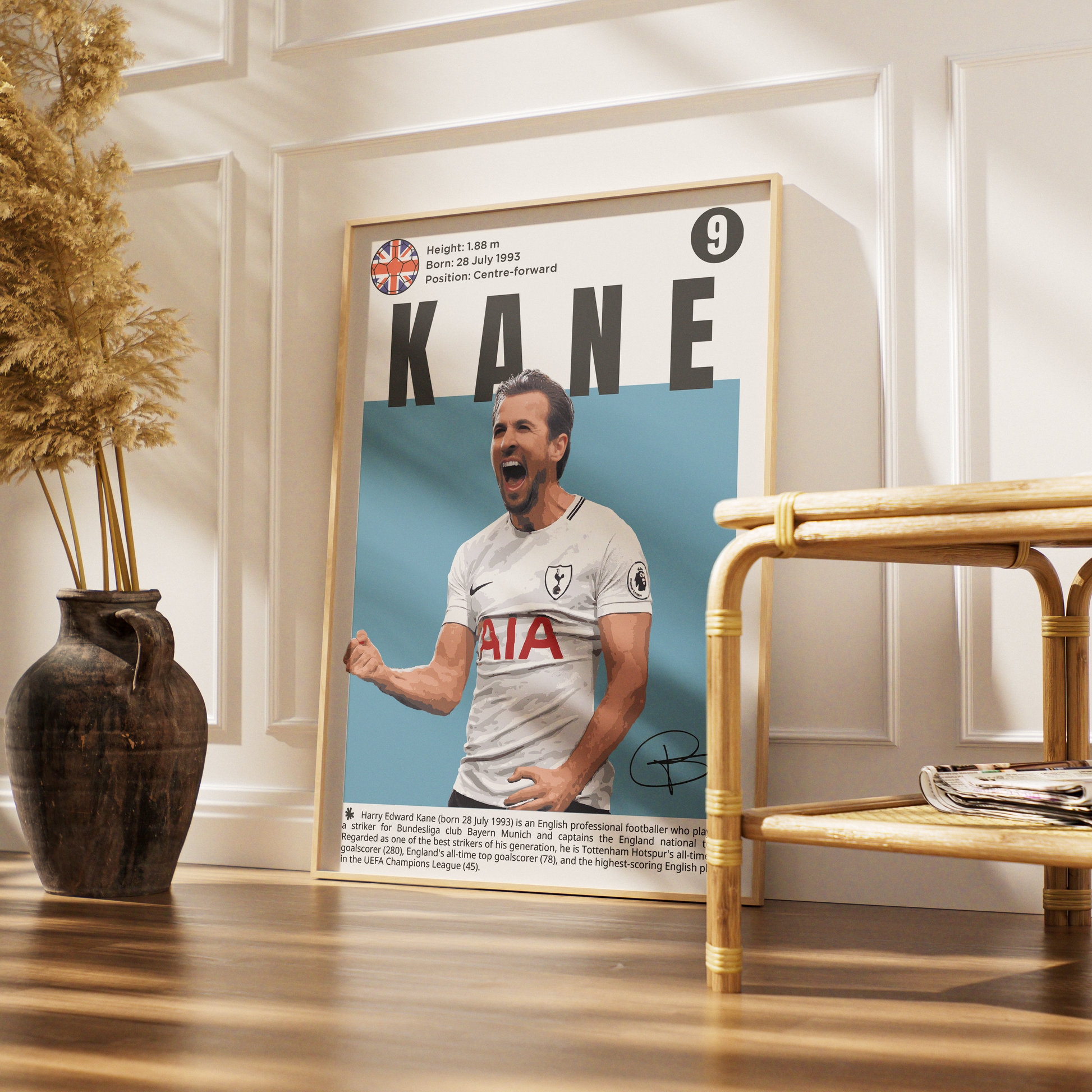 A framed poster featuring a portrait of footballer Kane in a white Tottenham jersey, displayed on a wooden floor in a styled interior with a vase of dried grass and a rattan side table.