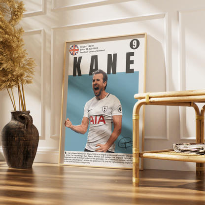 A framed poster featuring a portrait of footballer Kane in a white Tottenham jersey, displayed on a wooden floor in a styled interior with a vase of dried grass and a rattan side table.