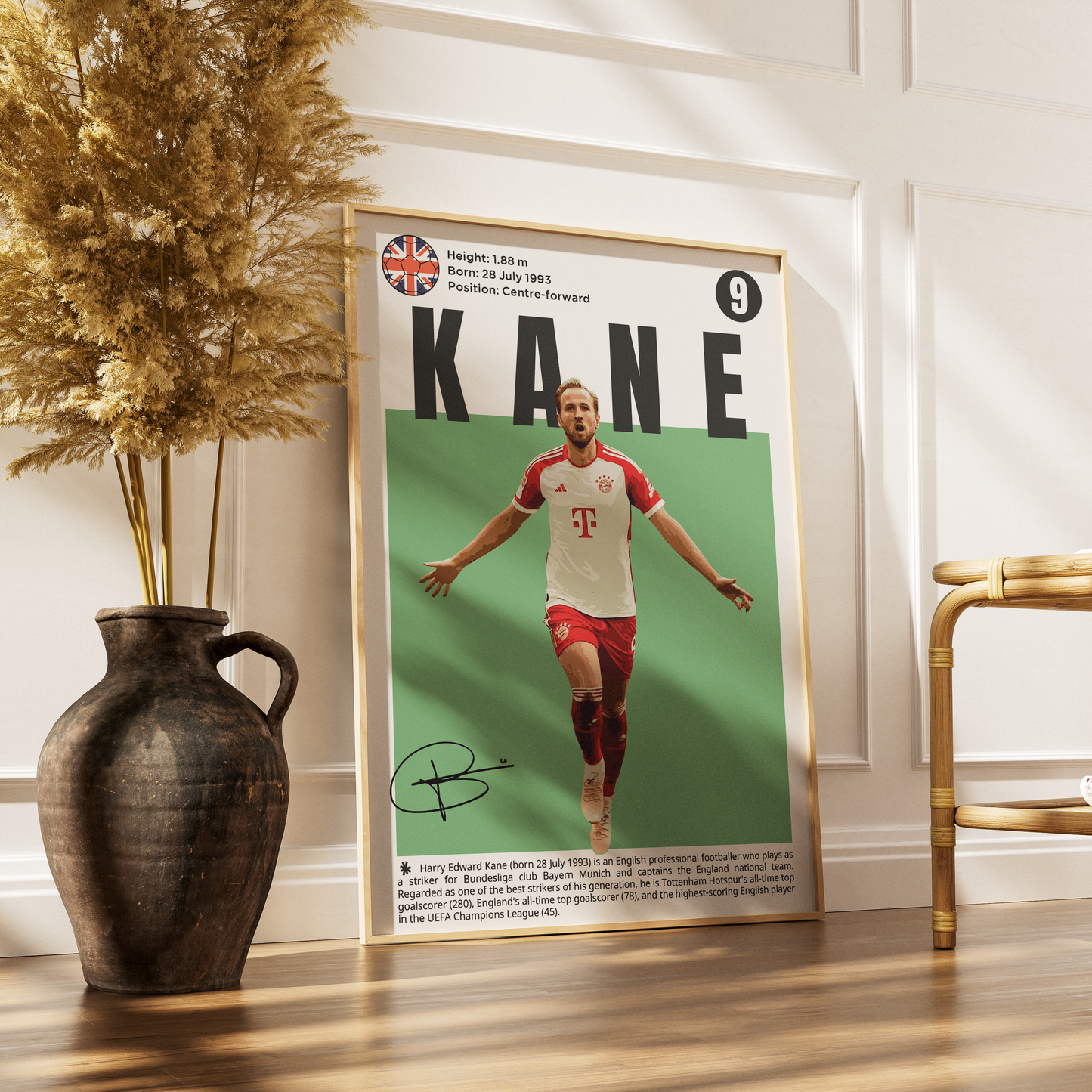 A framed vertical poster of soccer player Kane leaning against a white paneled wall on a wooden floor, styled with a dark ceramic vase of dried grass on the left and a gold chair on the right.
