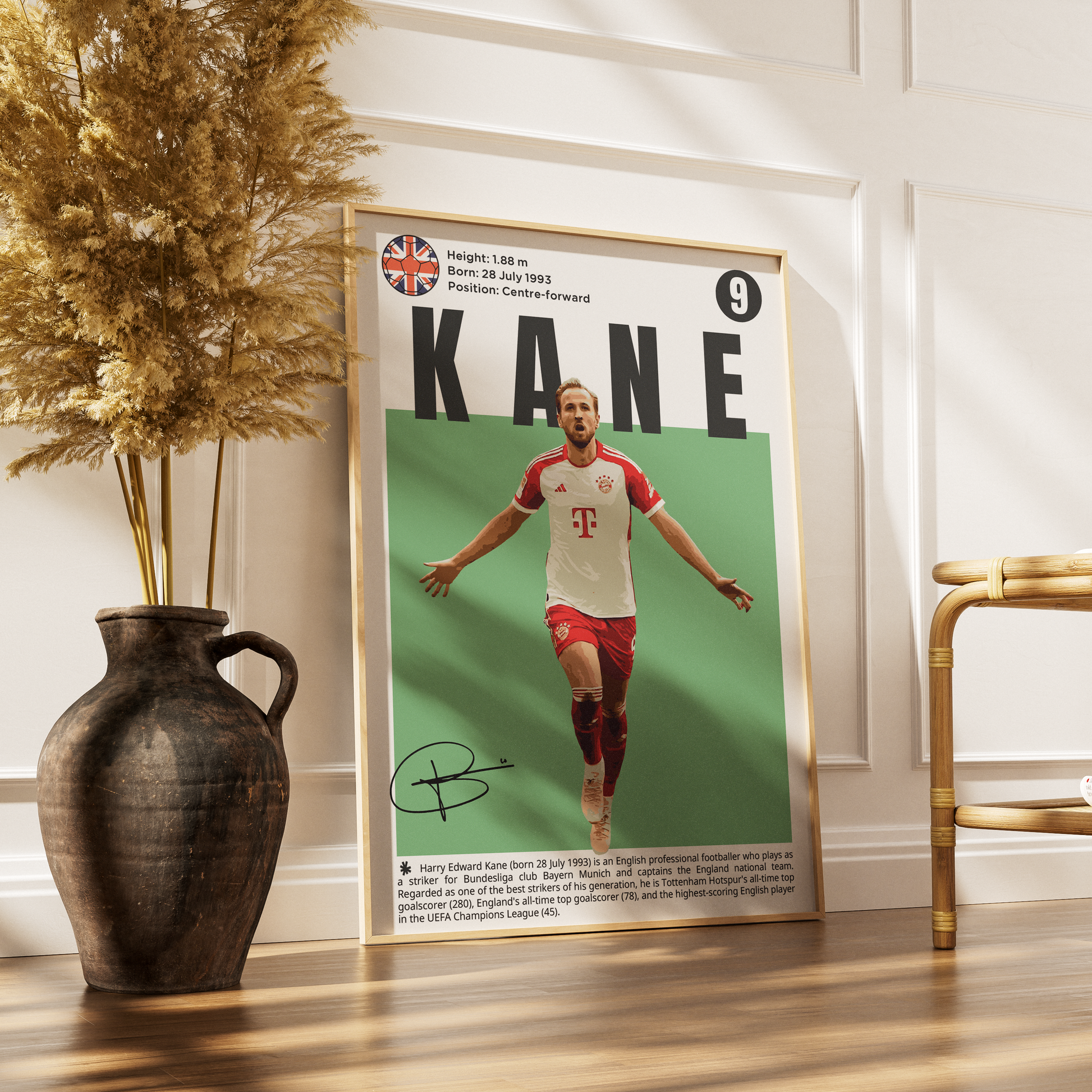 A framed vertical poster of soccer player Kane leaning against a white paneled wall on a wooden floor, styled with a dark ceramic vase of dried grass on the left and a gold chair on the right.