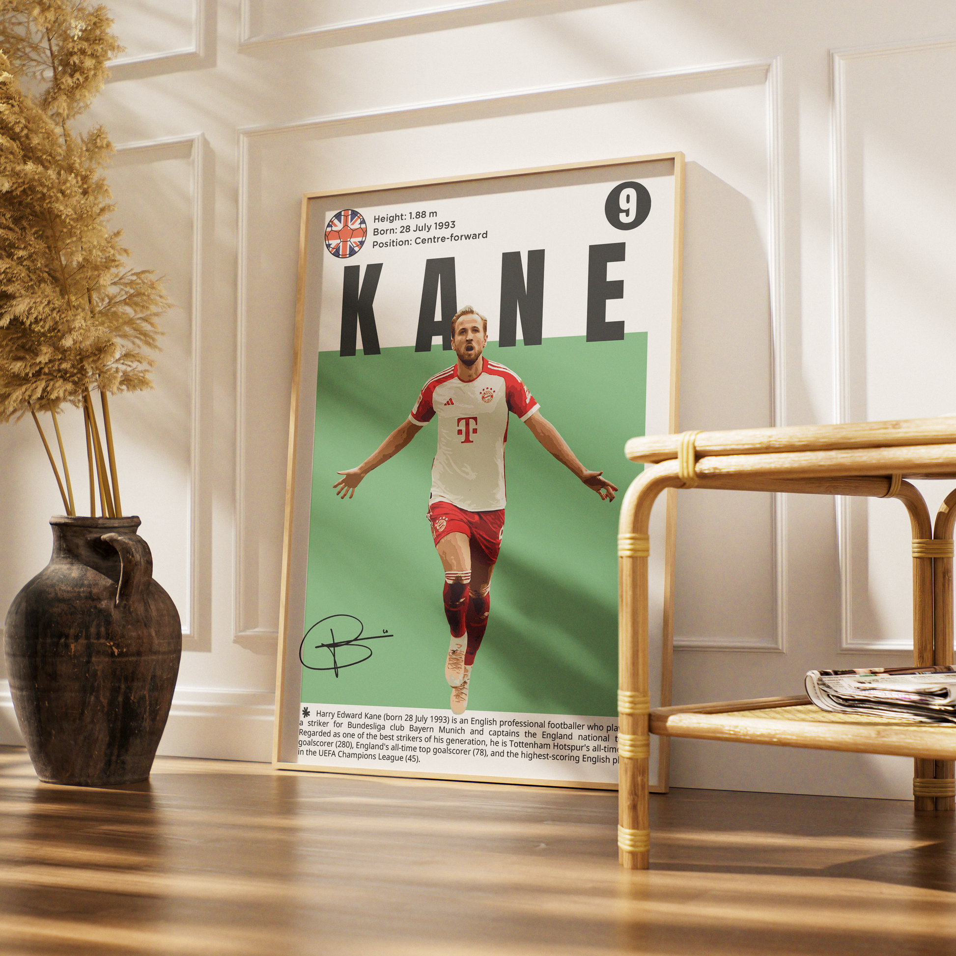 A framed soccer poster of Kane leaning against a white paneled wall in a styled room with a wooden floor, a dark vase with dried grass, and a rattan side table.