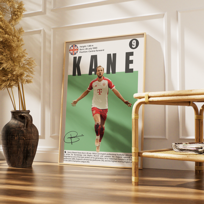 A framed soccer poster of Kane leaning against a white paneled wall in a styled room with a wooden floor, a dark vase with dried grass, and a rattan side table.