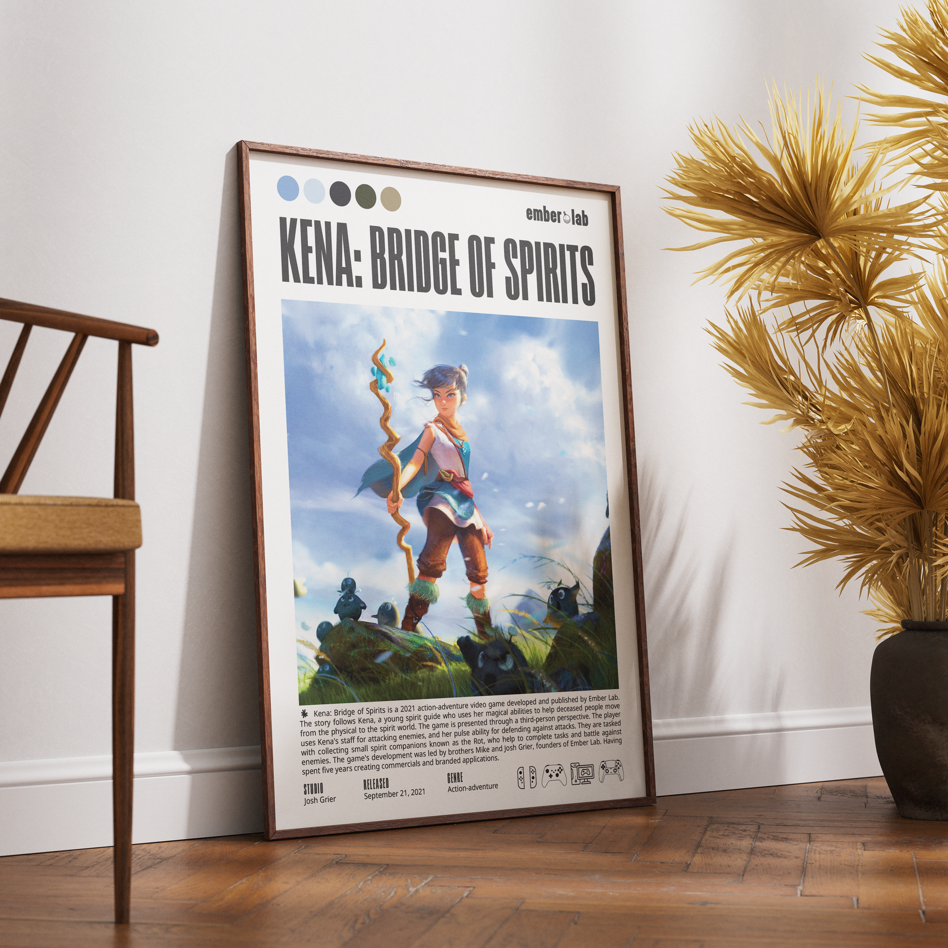 A framed Kena: Bridge of Spirits poster leaning against a white wall on a wooden floor, styled in a room with a wooden chair on the left and a potted palm on the right.