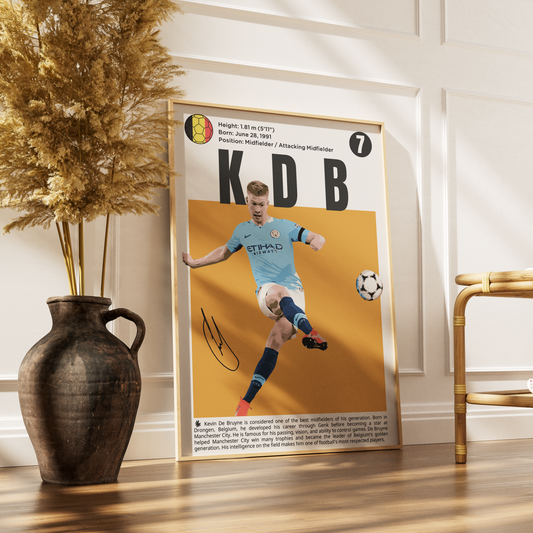 A framed football poster featuring Kevin De Bruyne in a light blue Manchester City kit, leaning against a white wall on a wooden floor next to a dark ceramic vase with dried grass and a gold chair.
