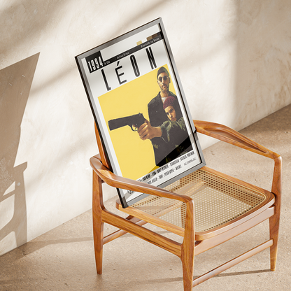 A framed movie poster for the film 'Léon' is displayed leaning on a mid-century modern wooden armchair with a cane seat, set against a textured beige plaster wall with soft diagonal shadows.