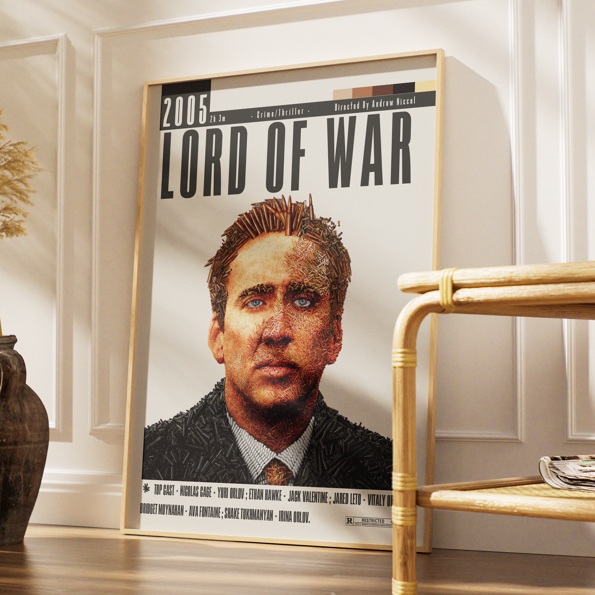 A framed movie poster for the 2005 film 'Lord of War' leaning against a white paneled wall in a styled interior setting with a bamboo side table and a dark vase.