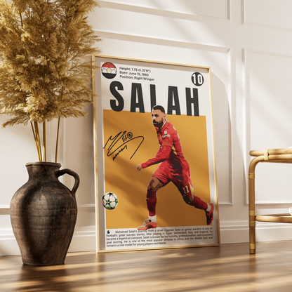 A framed sports poster of footballer Mohamed Salah leaning against a white paneled wall on a wooden floor, styled with a dark ceramic vase of dried grass on the left and a gold chair on the right.