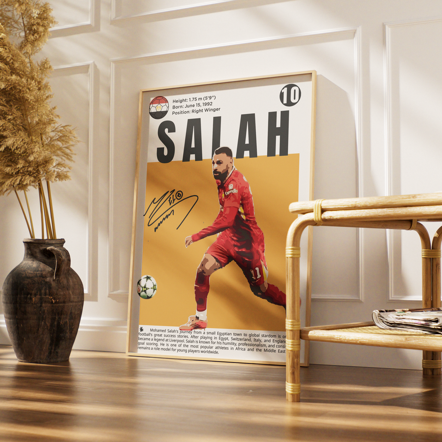 A framed personalized soccer poster of Mohamed Salah leaning against a white paneled wall in a modern room setting, next to a bamboo side table and a dark ceramic vase with dried grass.