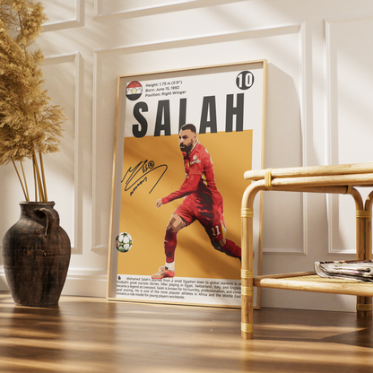 A framed personalized soccer poster of Mohamed Salah leaning against a white paneled wall in a modern room setting, next to a bamboo side table and a dark ceramic vase with dried grass.