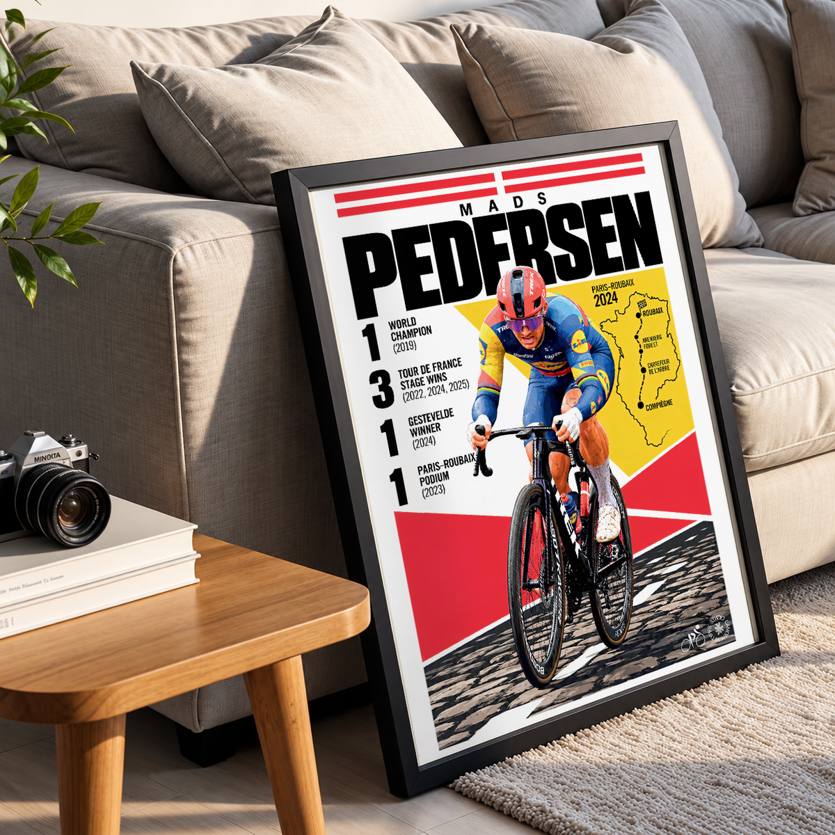 A framed cycling poster leaning against a beige sofa in a living room setting. The poster features a cyclist in a red and blue jersey on a road bike, with bold text reading 'Mads Pedersen' and race results.