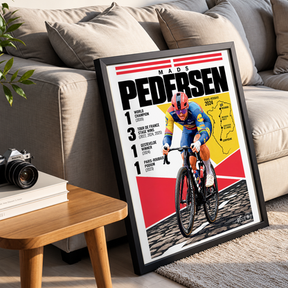 A framed cycling poster leaning against a beige sofa in a living room setting. The poster features a cyclist in a red and blue jersey on a road bike, with bold text reading 'Mads Pedersen' and race results.