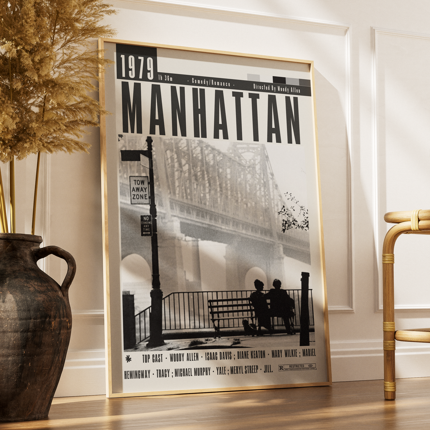 A framed black-and-white poster titled "1979 Manhattan" leaning against a white wall on a wood floor, styled in a room with a large dark vase and dried grass on the left and part of a bamboo chair on the right.