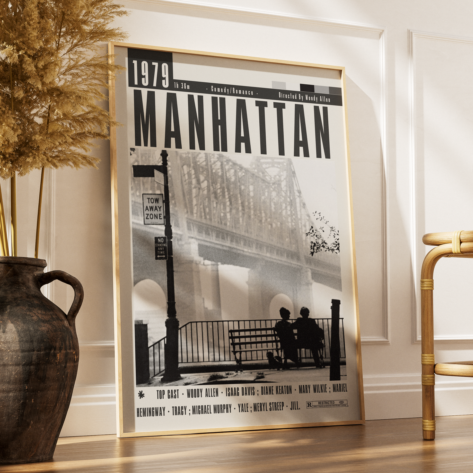 A framed black-and-white poster titled "1979 Manhattan" leaning against a white wall on a wood floor, styled in a room with a large dark vase and dried grass on the left and part of a bamboo chair on the right.