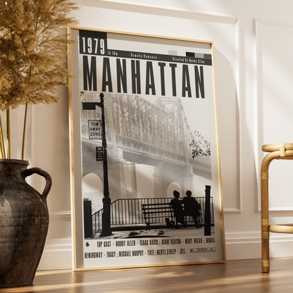 A framed black-and-white poster titled "1979 Manhattan" leaning against a white wall on a wood floor, styled in a room with a large dark vase and dried grass on the left and part of a bamboo chair on the right.