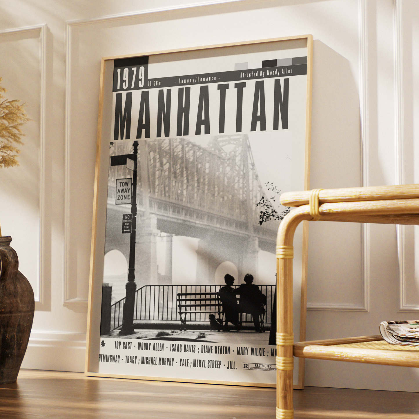 A framed black-and-white Manhattan magazine cover print leaning against a white paneled wall in a styled interior, with a bamboo side table and decorative vase nearby.