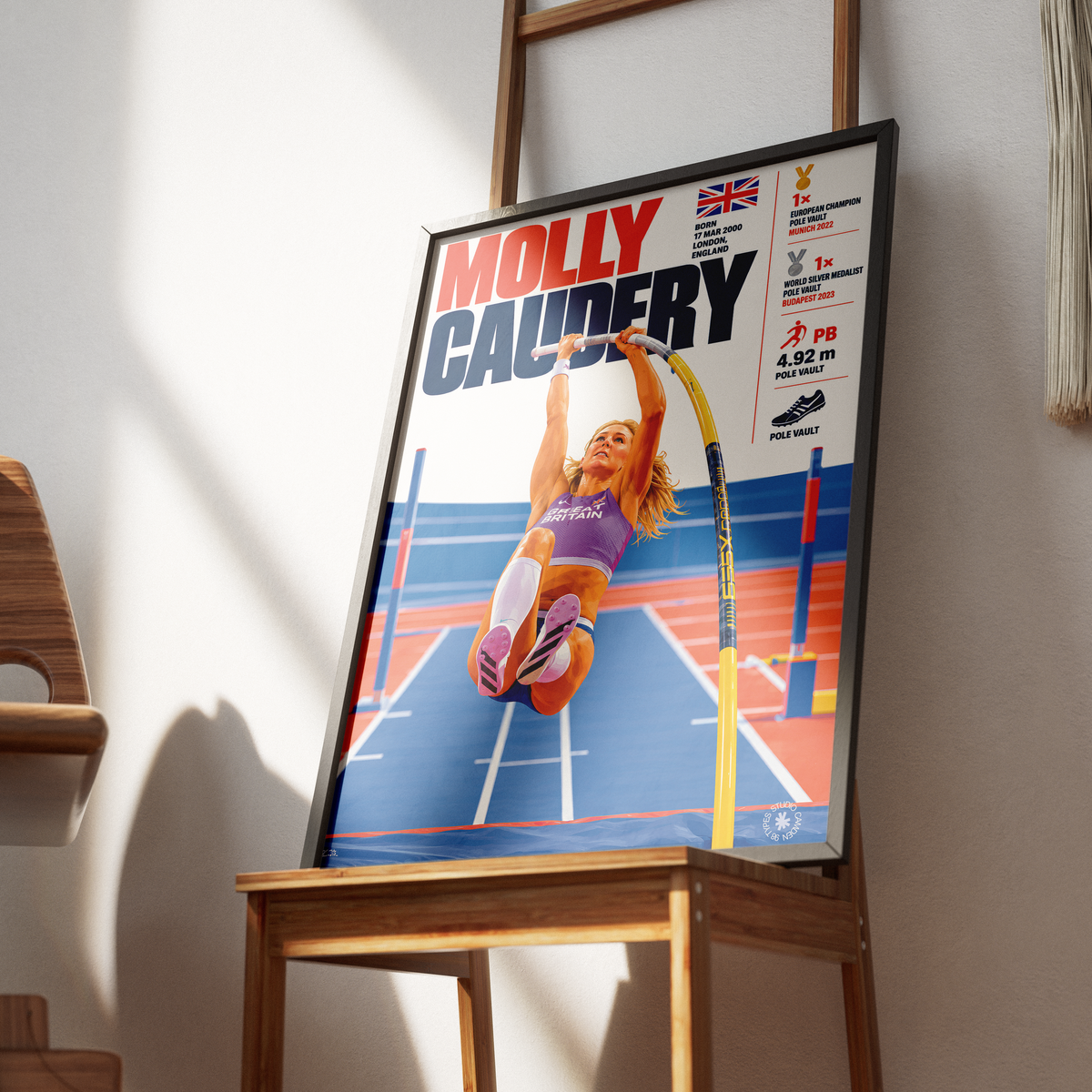 A framed poster of athlete Molly Caudery leaning against a wall on a wooden stand in a sunlit room. The poster features a photo of Caudery jumping on a high jump bar, with text including 'MOLLY CAUDERY' and athletic achievement details.