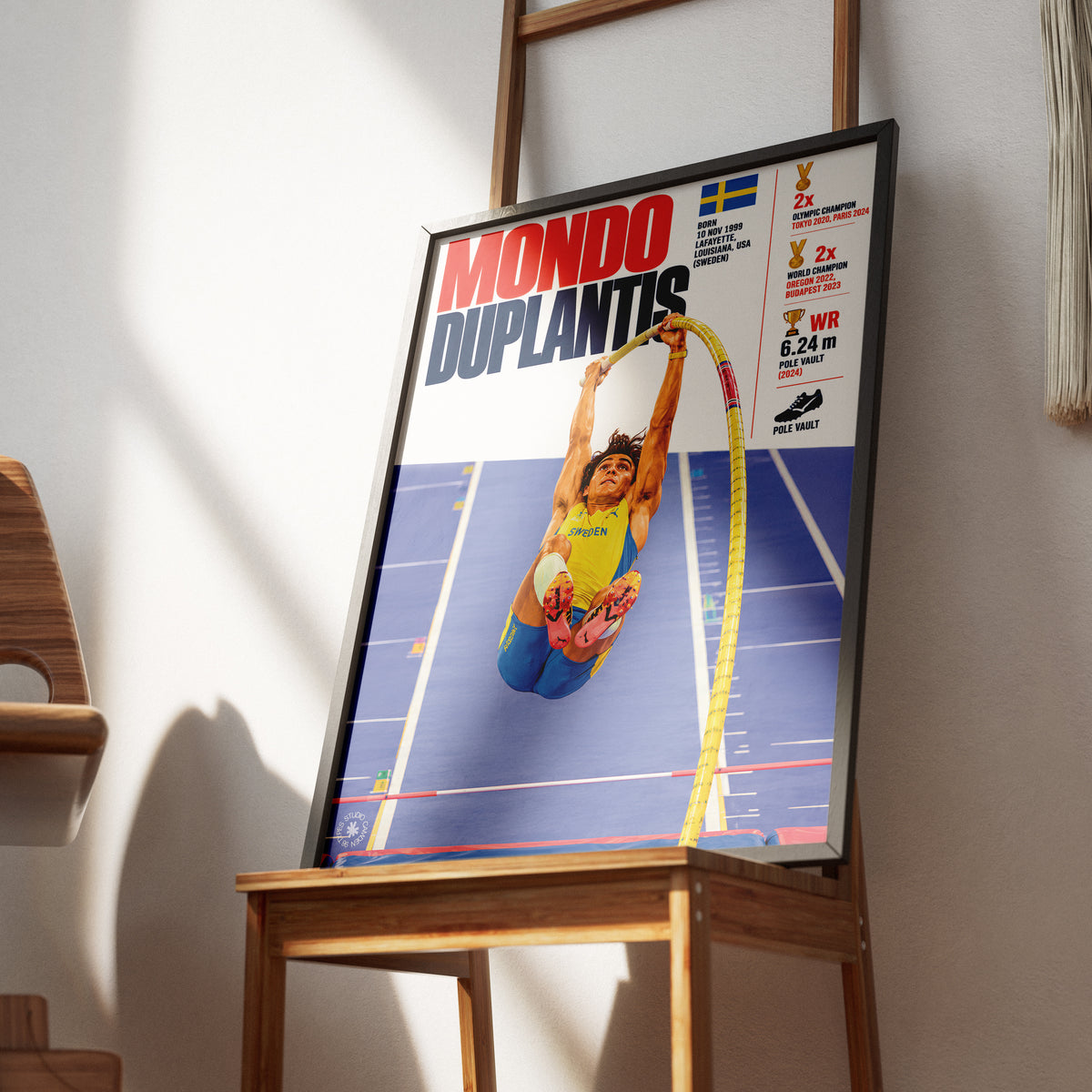 A framed Mondo Duplantis poster leaning on a wooden chair in a sunlit room. The poster features athlete Kippen Andersen jumping the pole, with Swedish flag graphics and competition results on the right side.
