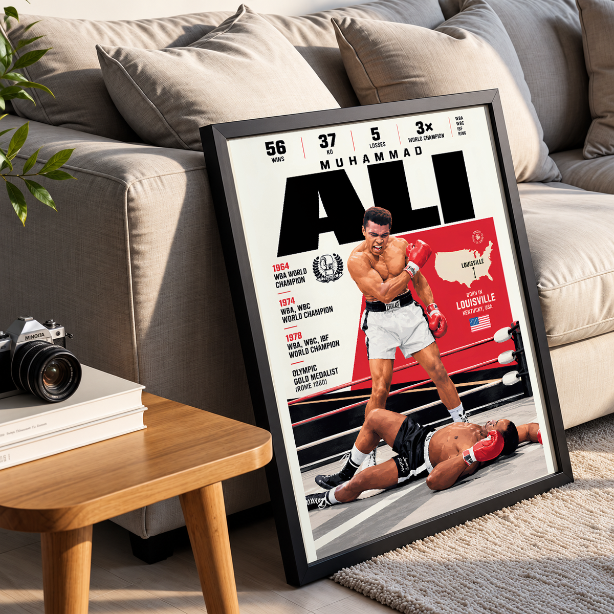A framed poster of Muhammad Ali leaning against a beige sofa in a living room setting, with a wooden side table and camera nearby.