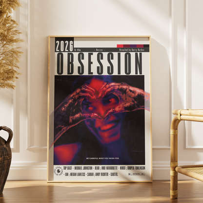 A framed poster for the movie 'Obsession' leaning against a cream-colored paneled wall on a wooden floor, with a dried pampas grass vase on the left and a rattan chair on the right.