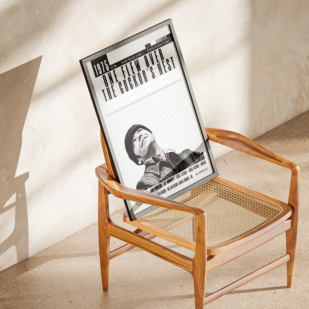 A framed black-and-white poster leaning against a wooden armchair in a sunlit room with a textured beige wall and floor.
